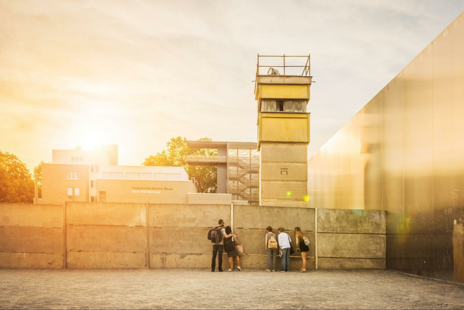 A bleak, monotone photo depicting visitors looking through a slat in the concrete of a former watchtower at the Berlin Wall Memorial. 