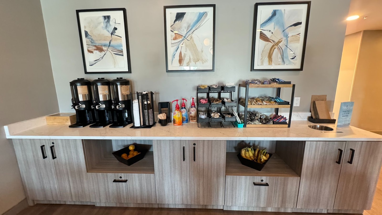 Three pieces of artwork and a large breakfast bar with coffee carafes, creamers and organizers with fruit and granola bars at at Hyatt Studios Mobile Tillman's Corner