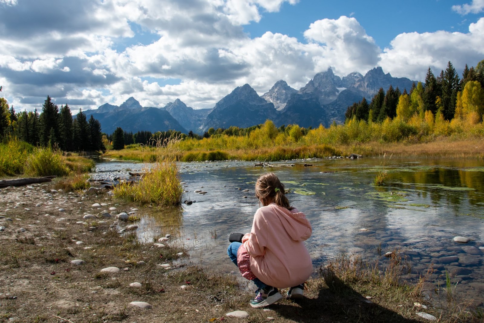 A young girl playing with the water along the riverbank surrounded by mountains and fall colored trees