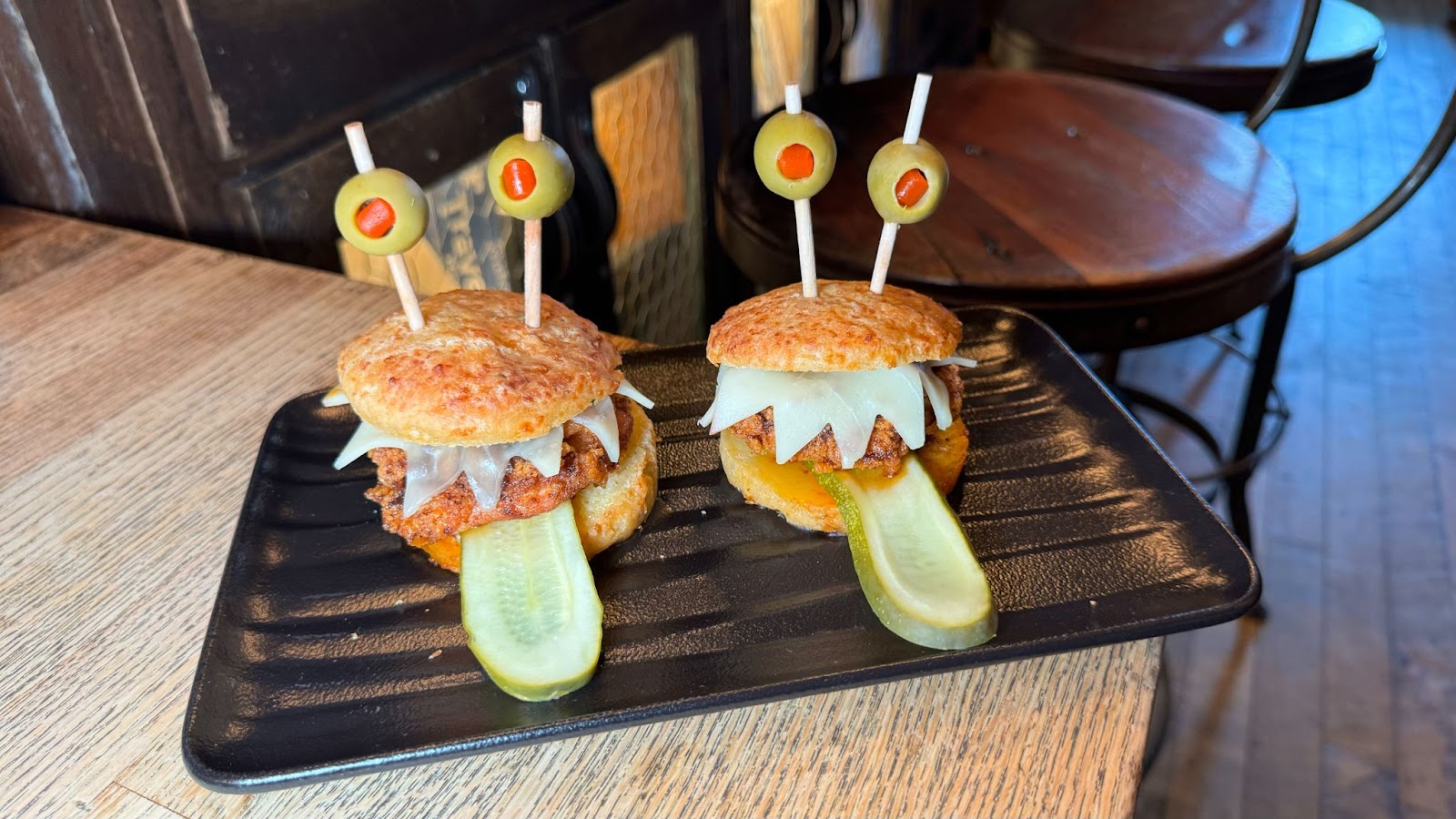 Fried chicken sliders on top of a black plate resting on a wooden table at a Halloween bar in Orlando known as Jock Lindsey's Halloween Hangar Bar.
