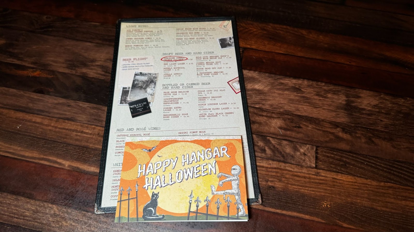 A menu along with a card saying Happy Hangar Halloween on a wooden table at a Halloween-themed bar in Orlando.