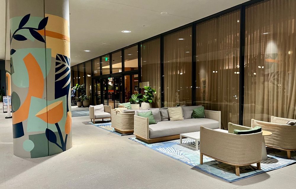 Seating area outside the hotel with upholstered couches, chairs and throw pillows against the hotel windows at night. A mural covers a large pillar.
