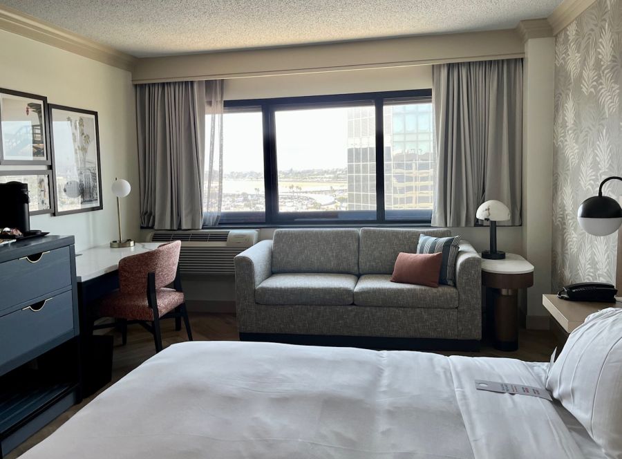 A Preferred King room, decorated in cream and gray. In the forefront is a bed with gray linens, with gray couch below a window behind it, plus a desk, chair, night table, side table and lamps.
