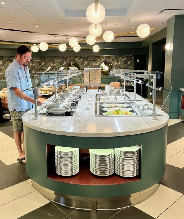 A man in a blue button-down shirt and gray shorts stands at a buffet with serving dishes on one side and sunken serving dishes on the other. Plates are stacked in a cut-out at the front.