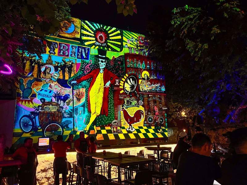 A colorful mural on the back wall of a bar, featuring a large skeleton and other Mexican culture objects.