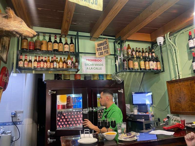 Inside of a bar, with alcohol bottled lined on the wall,  a cabinet with drinks, a waiter behind the bar, and glasses of wine and plates of food on top of the bar