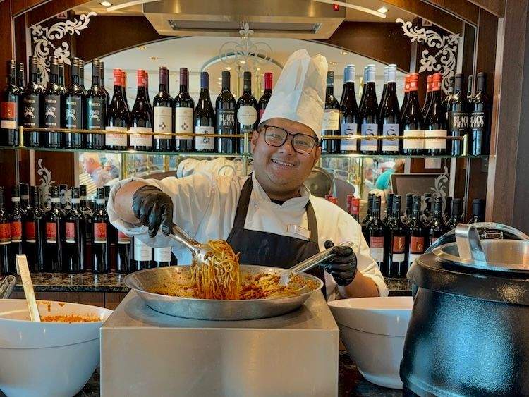 Crew member serves up spaghetti Bolognese in front of bar with wine bottles in the MS George Eliot’s dining room.