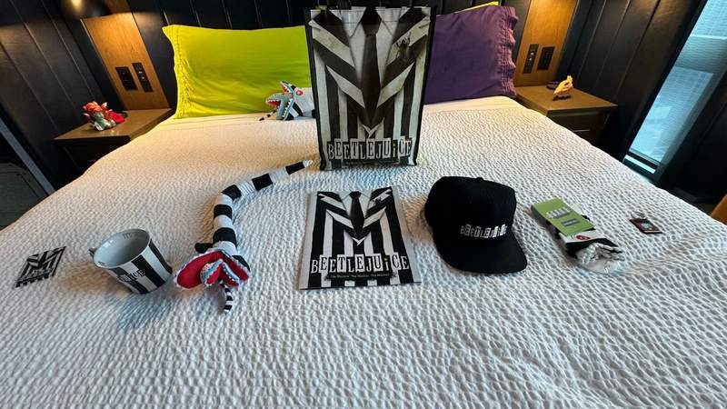 white bed with beetlejuice magnet, mug, plush, program, hat, socks and keychain inside the Beetlejuice suite.