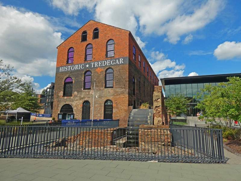 There story brick building with a water wheel in front and modern glass building on right  side.