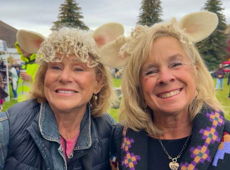 Two women wearing whimsical lamb ear and curly wool headbands enjoying the Folklife Fair with food and vendor booths in the background.