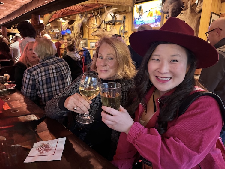 Two women seated at the bar of the historic Pioneer Saloon in Ketchum, raising glasses in a celebratory toast during a girls’ getaway