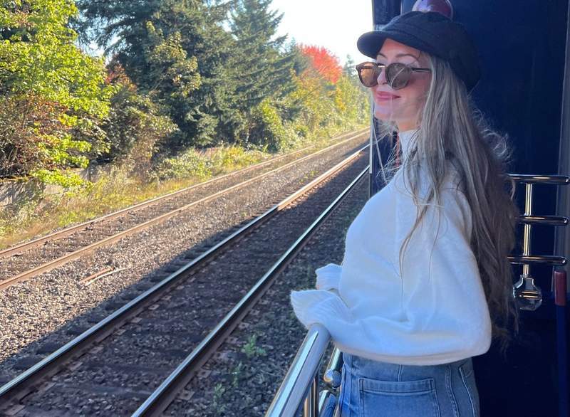 Woman dressed in warm sweater, jeans, and a cap enjoys the scenery on the outdoor train viewing platform.