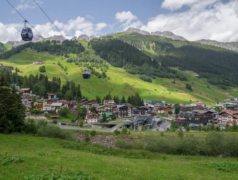 The village of St. Anton am Arlberg in the summer showing the gondola going up the mountain and the village beneath, surrounded by the peaks of the Austrian Alps.