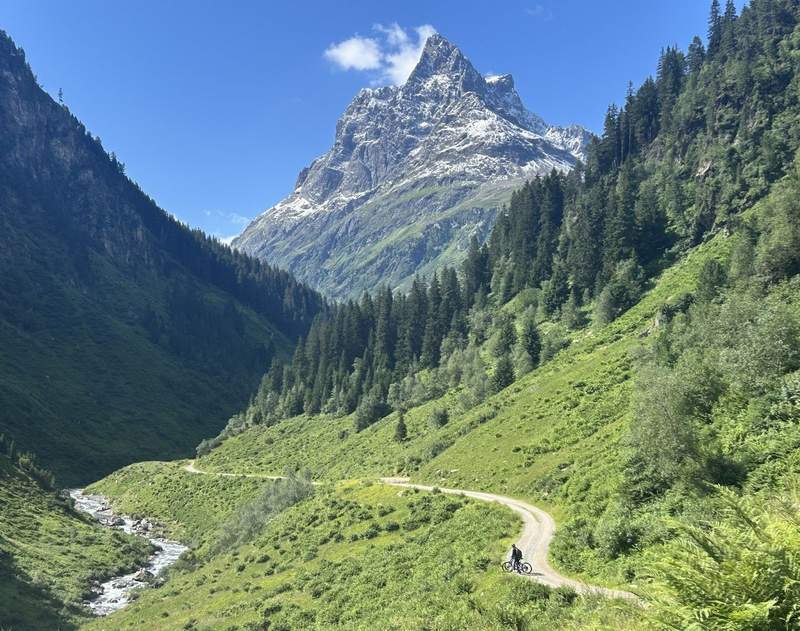 An e-biker, looking small compared to the high mountain peaks, on a trail in the  Verwalltal, a high-alpine wilderness area in the Arlberg region of the Austrian Alps. 