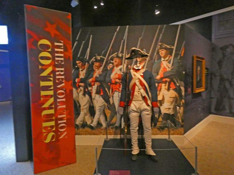 A museum display with a troup of Continental soldiers in background , a Revolutionary War military uniform in foreground and a sign saying 