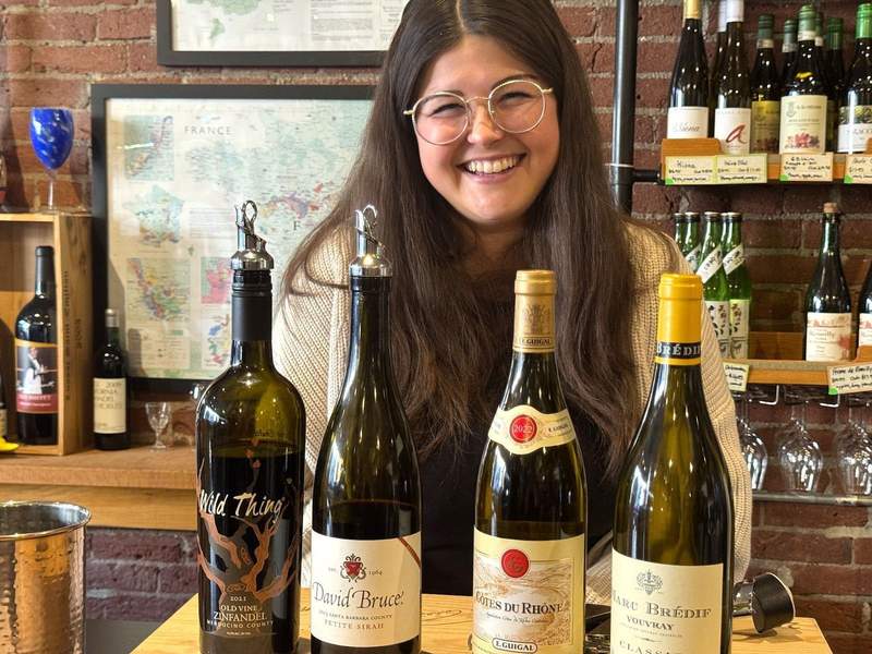 Happy woman serves wine at a bar. Four wine bottles in front of her and the shelves behind her has wine bottles.