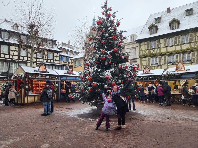A mother and daughter standing in front of a Christmas tree in Colmar with snow dusting the roof and treetops