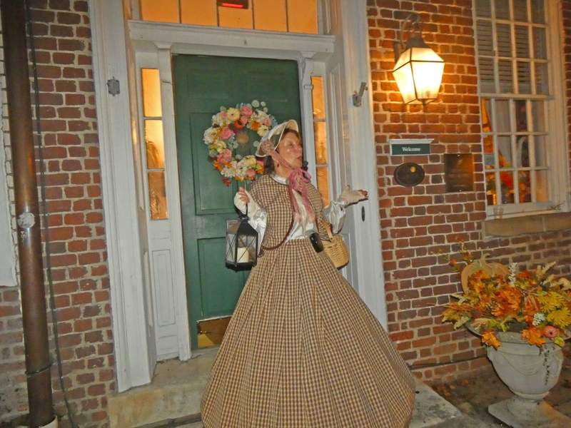 A woman in colonial costume in front of a green door on a brick building.