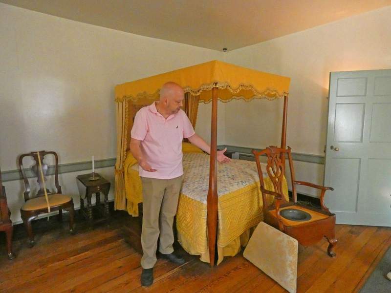 A man in front of a colonial four poster bed with a chamber pot chair in front.