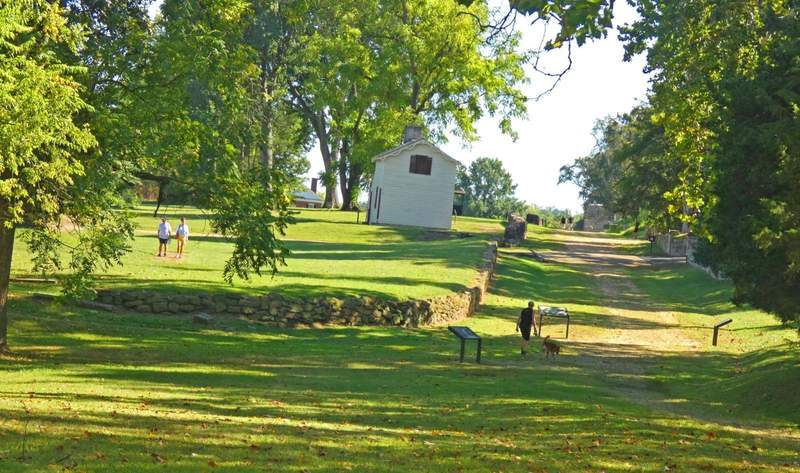 A green landscape with a stone fence and white building  and several people  walking.