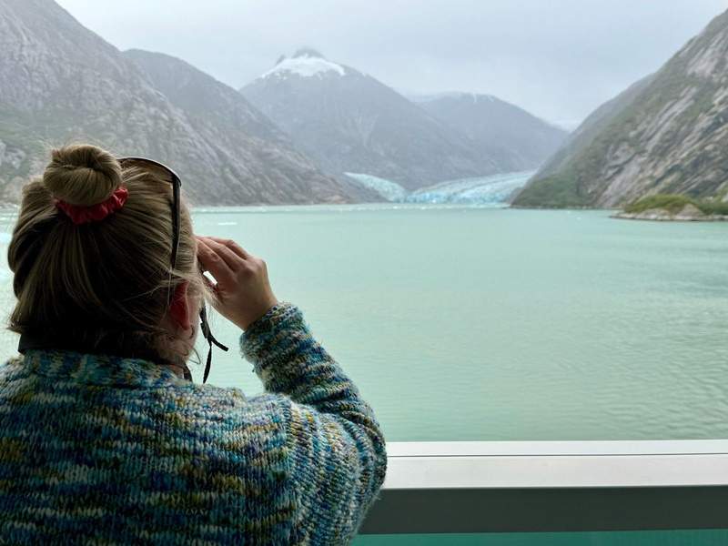 Woman looks through binoculars at the Dawes Glacier and mountains from an Infinite Veranda on the Celebrity Edge