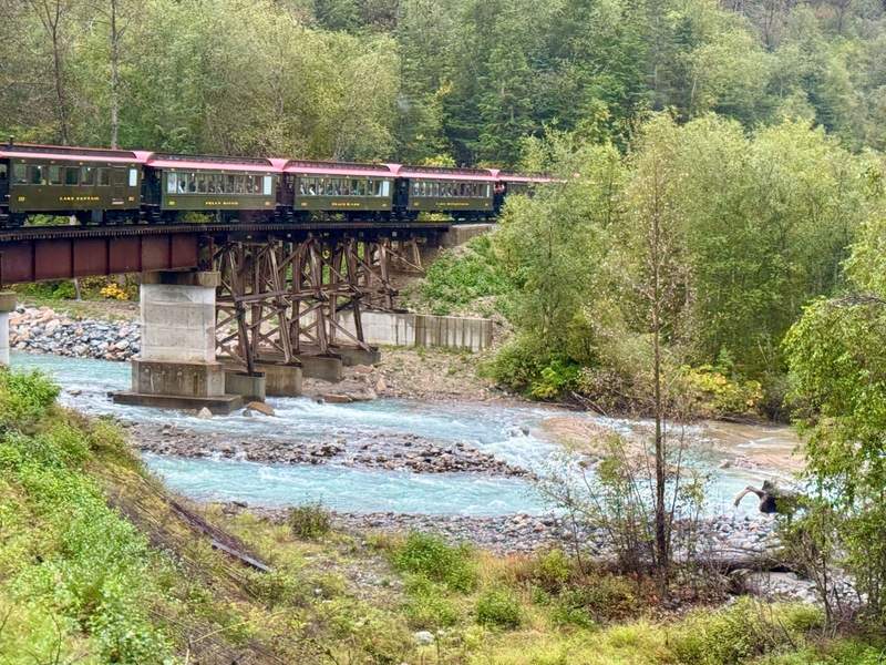 The White Pass Scenic Railway crosses a bridge over rushing water. Trees surround the tracks.