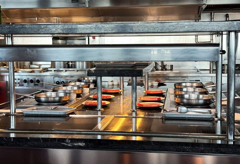 A stainless steel kitchen with six skillets, three on each side with everything to cook a steak.
