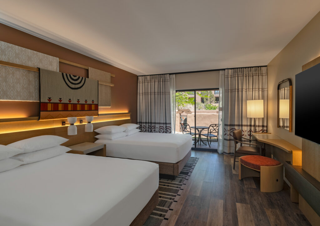 A remodeled guest room at the Hyatt Regency Tamaya Resort & Spa featuring two queen beds, warm earth-tone décor, Pueblo-inspired textiles, and a private patio with outdoor seating. Natural light fills the room through sliding glass doors overlooking the resort grounds.