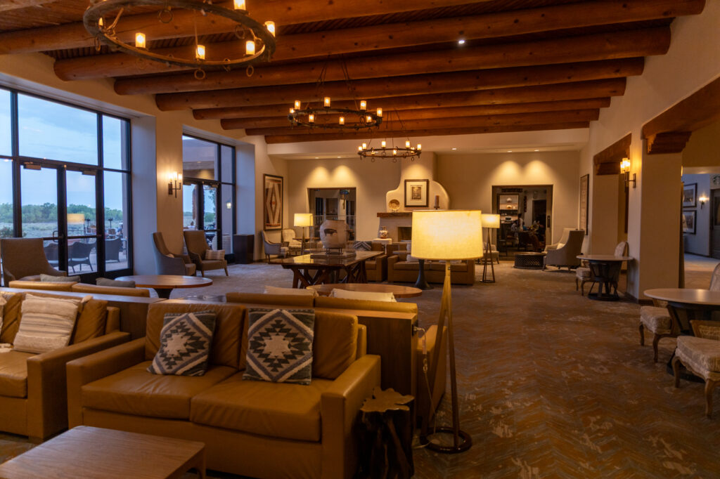 The Rio Grande Lounge at the Hyatt Regency Tamaya Resort & Spa features leather sofas, Pueblo-patterned pillows, and warm lighting beneath viga-style wooden beams. Large windows reveal the outdoor patio and the Rio Grande Valley beyond, creating a cozy Southwestern ambiance.