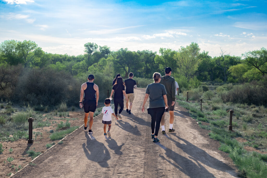 A group of guests, including a child, walk along a dirt nature trail at the Hyatt Regency Tamaya Resort & Spa. Morning sunlight casts long shadows as they head toward the cottonwood bosque near the Rio Grande, surrounded by desert brush and greenery.