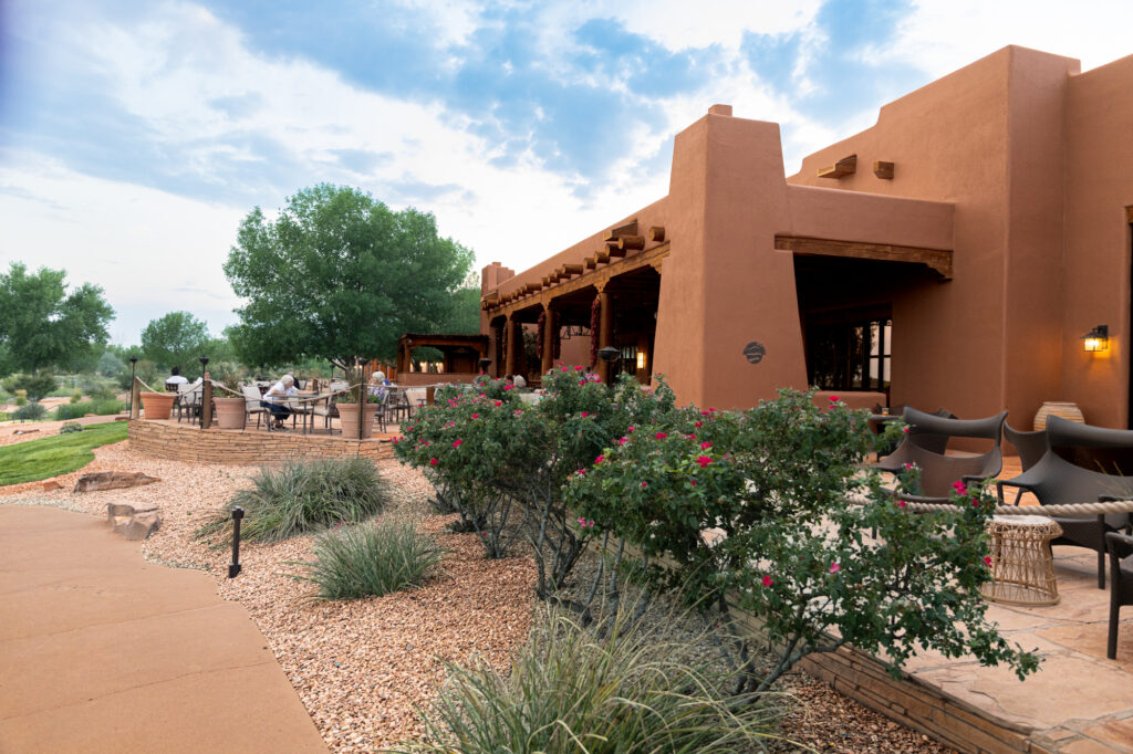 Guests relax on the outdoor patio of the Rio Grande Lounge at the Hyatt Regency Tamaya Resort & Spa. The adobe-style building features wooden vigas, warm earth tones, and desert landscaping with flowering shrubs and stone pathways overlooking the Rio Grande Valley.