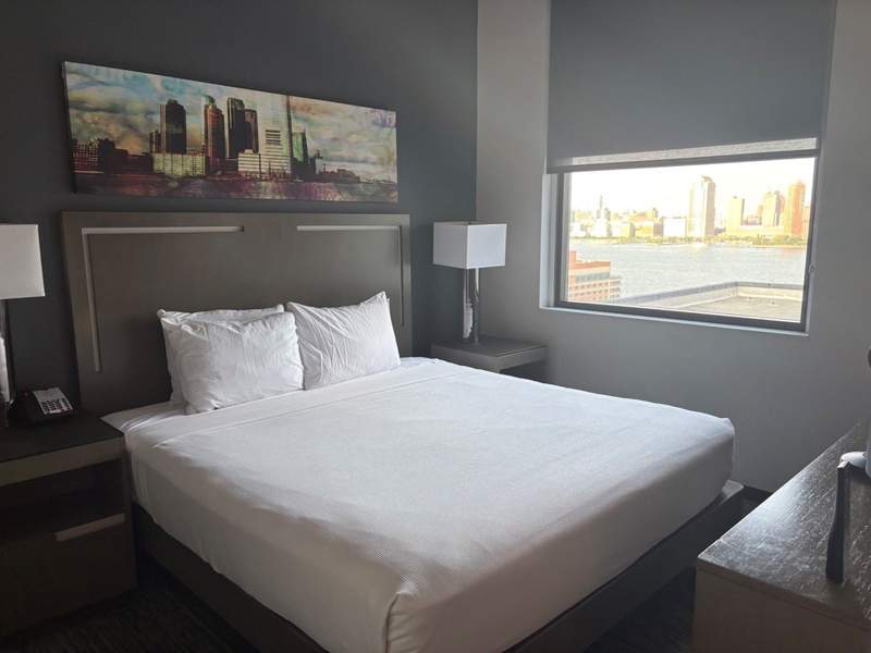 A white bed with modern decor and a window view of the city skyline.