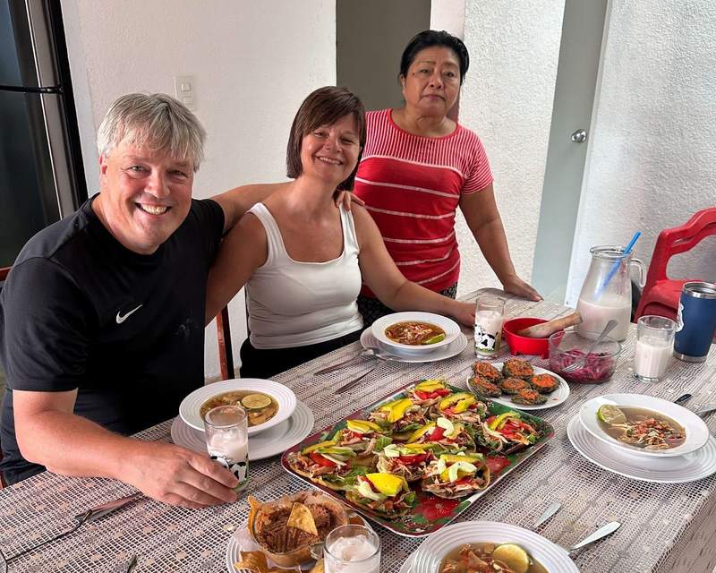 A couple of tourists sitting at the table with a local woman standing beside them. Typical dishes can be seen on the table.