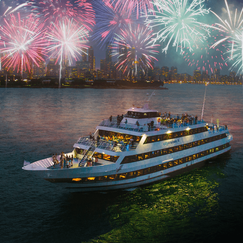 View of a multi-story enclosed boat with crowds of people, on the water in front of a cityscape at night with fireworks overhead.