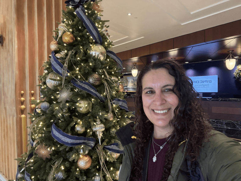 A woman with brown hir in front of a Christmas tree decorated with gold and blue. 
