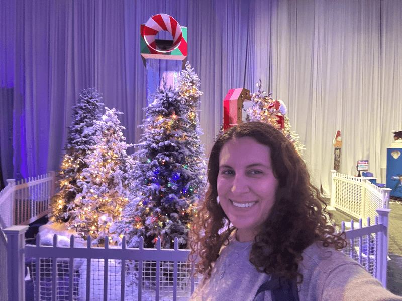 A woman with brown hair in front of Christmas trees covered in faux snow. 