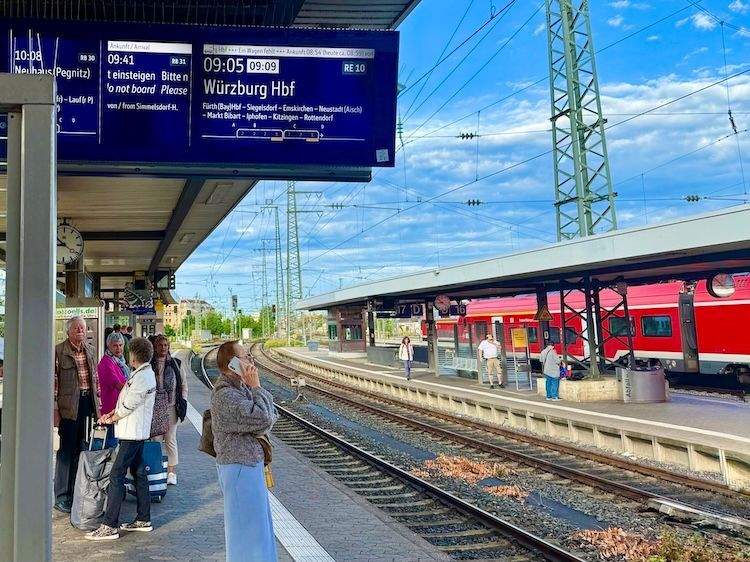 View of Nuremberg train platform with travelers standing and walking, including a woman on her phone, as everyone readies for train to arrive.