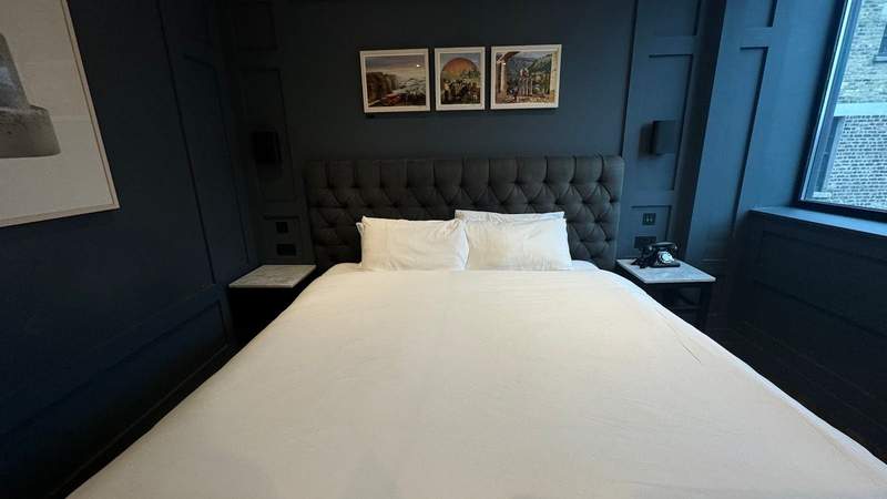 White bed with pillows inside a navy blue room next to a window with three pieces of artwork at The Dean Dublin.