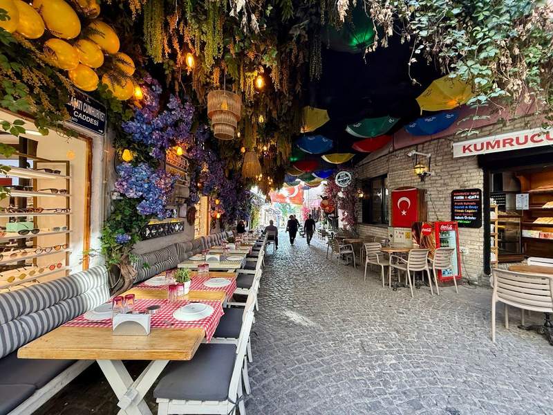 Lovely cobblestone street lined with colorful restaurant  tables and chairs and covered above by trees, plants, and colorful lights and decorations.