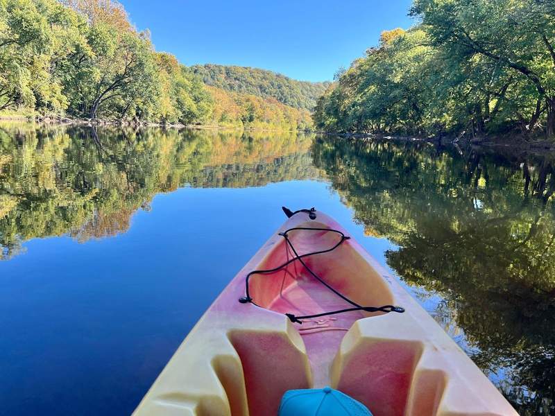 Tip of a kayak pointing down the James River with Blue Ridge Mountain foliage on the riverbank. 