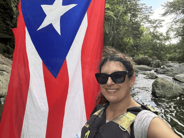 Women in front of the Puerto Rican flag in a river. She is wearing a life vest and sunglasses.