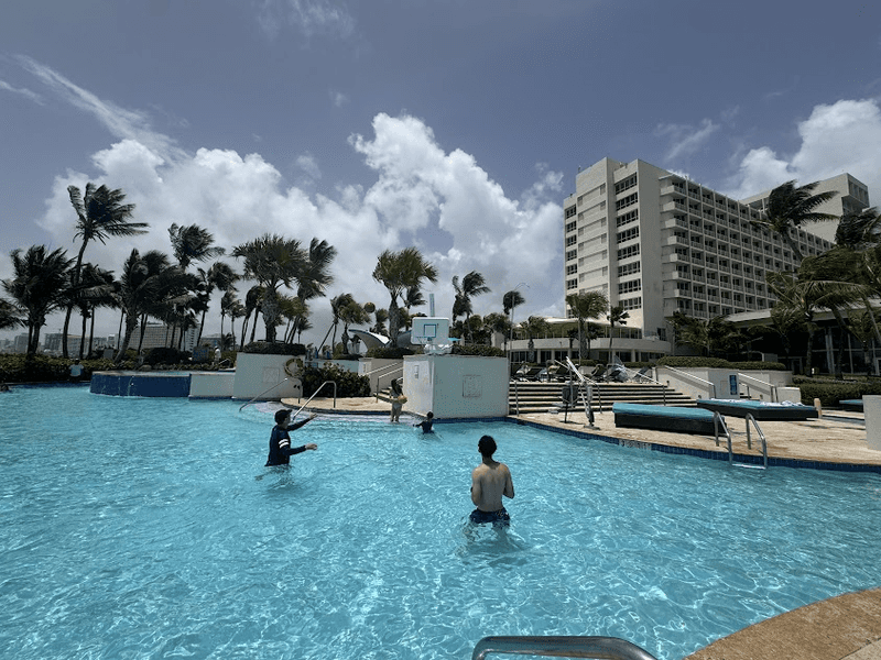 A man and children playing basketball in a poo. There are plan trees and a hotel in the background. 