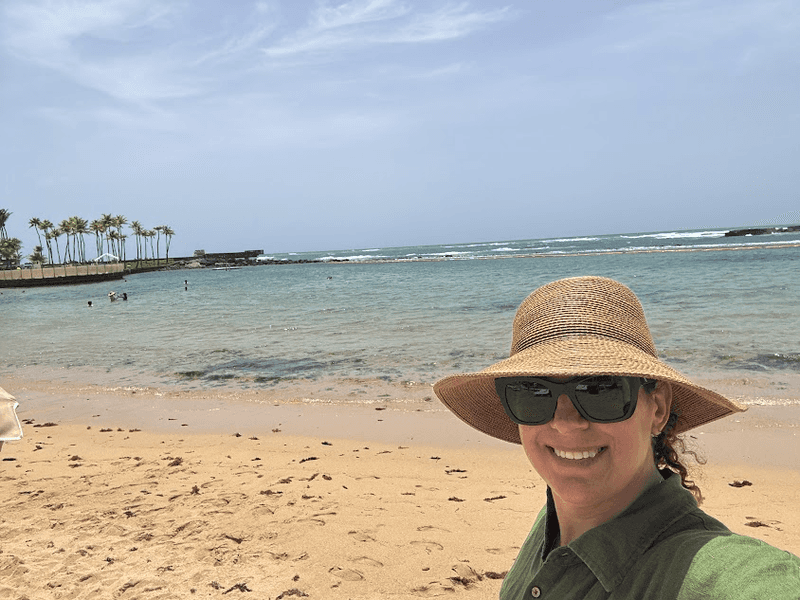 Woman in a hat and sunglasses on a secluded beach with palm trees in the background.