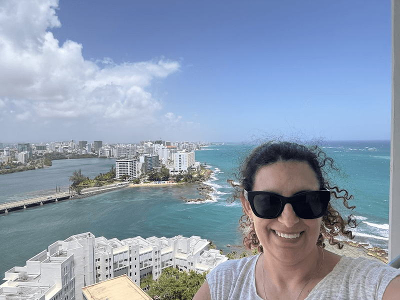 Woman in sunglasses with a view of an ocean and an island with buildings in the background. 