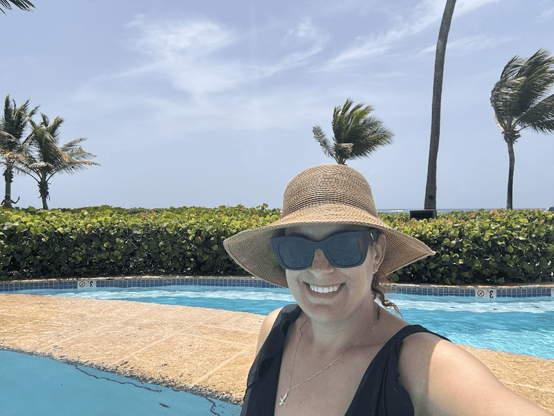 A woman in a hat and sunglasses by the pool. There are bushes and palm trees in the background. 