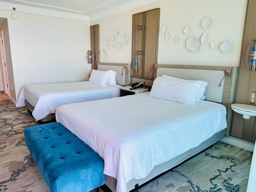 View of a hotel room with two beds with white linens. One bed has a wide blue ottoman at the end.