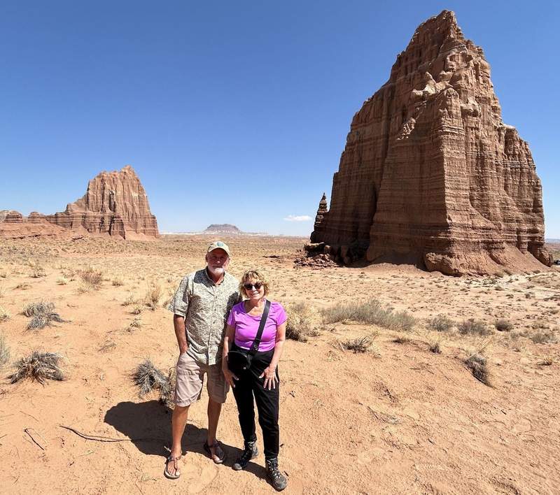 The author in a lavender t-shirt with a bearded tour guide from Water Pocket Adventures stand on the brown desert with two large reddish rocks poking up in the distance in an open landscape.