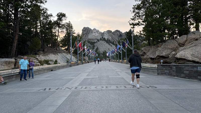 sunset photo of people walking down the avenue of flags with different flags hanging up on each side in front of Mount Rushmore. 