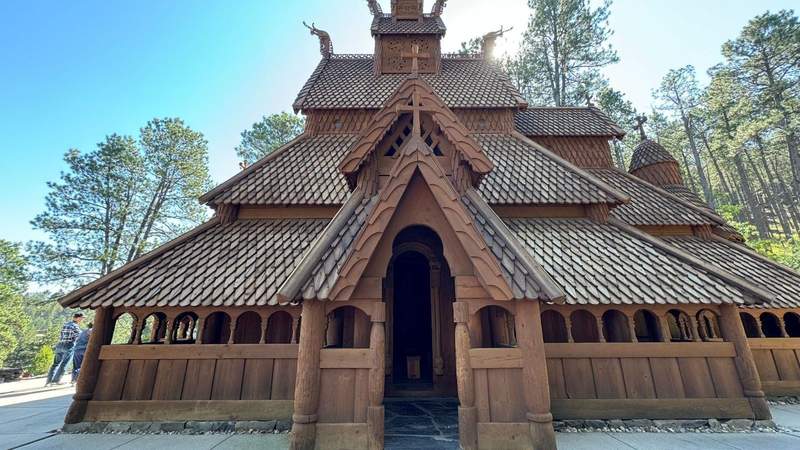Wooden Norwegian inspired church with three roofs and oval shaped tiles infront of trees in South Dakota.