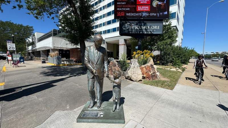 Bronze life-size statue of John F. Kennedy next to a small child on the corner outside of a hotel. 